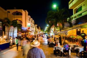 Por la noche, una calle animada y colorida, con gente paseando y cenando al aire libre en restaurantes cercanos a los vibrantes hoteles de México; palmeras y luces brillantes iluminan el ambiente, perfecto para conferencias y reuniones.