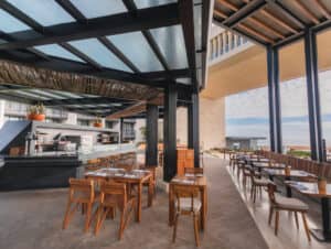 Un restaurante moderno al aire libre en los complejos turísticos de México, con mesas y sillas de madera, una zona de cocina central y grandes ventanales con vistas al océano, perfecto para reuniones o eventos de conferencias bajo un cielo parcialmente nublado.