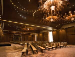 Una sala de conferencias amplia y elegante en uno de los mejores complejos turísticos de México, con filas de sillas beige, un pequeño escenario a la izquierda, paredes de madera e iluminación tenue: perfecta para eventos de conferencias memorables o reuniones exclusivas.