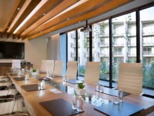 Una moderna sala de conferencias en uno de los resorts más exclusivos de México, equipada con una larga mesa de cristal, sillas de cuero blanco, blocs de notas y botellas de agua. Sus amplios ventanales ofrecen vistas a balcones y zonas verdes, ideales para reuniones o eventos.