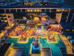 Vista aérea de una moderna zona de piscina al aire libre iluminada por la noche, donde la gente se reúne para eventos y conferencias rodeada de luces brillantes, piscinas y decoración, todo ello con el telón de fondo de un gran hotel de varias plantas en México.