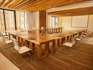 Una moderna sala de conferencias en un resort mexicano, con mesas de madera en forma de U, sillas blancas, vasos de agua de cristal, blocs de notas, un proyector y una gran pantalla en blanco: ideal para reuniones y eventos. Los paneles de madera en el techo y los grandes ventanales completan el espacio.