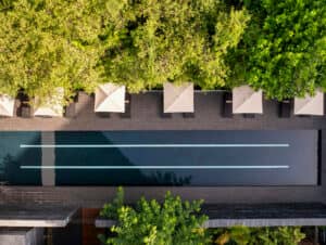 Vista aérea de una piscina rectangular con dos carriles de natación, rodeada de baldosas de piedra oscura, tumbonas, sombrillas beige y frondosos árboles verdes; perfecta para relajarse entre eventos de conferencias en los mejores complejos turísticos de México.