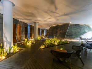 Moderna sala de estar interior con sillas y mesas de mimbre, pasarelas de madera, plantas decorativas y paredes de piedra: ideal para conferencias o reuniones en resorts de lujo en México. La iluminación tenue realza la atmósfera serena y sofisticada.