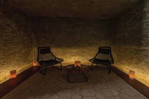 Una habitación con iluminación tenue, dos sillas negras de mimbre, una mesa baja de madera, paredes de piedra y cuatro luces que parecen velas crean una atmósfera relajante, similar a la de un spa, perfecta para reuniones íntimas o para relajarse en los complejos turísticos de México.