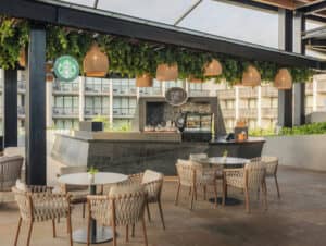 Zona de estar al aire libre con mesas redondas y sillas de mimbre frente a un moderno mostrador de Starbucks bajo una pérgola, perfecta para reuniones informales o eventos de conferencias en los complejos turísticos de México; se pueden ver la vitrina y la barra de café espresso.