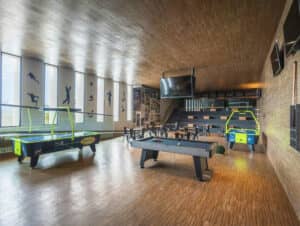 Una espaciosa sala de juegos en un resort mexicano con mesas de air hockey, mesa de billar, mesas y sillas, grandes ventanales con siluetas deportivas, arte mural, una escalera y un televisor montado en la pared. Ideal para reuniones o eventos. Los pisos y el techo de madera completan la decoración.