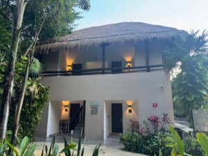 Un edificio de dos plantas con techo de paja, perteneciente a un complejo turístico, está rodeado de plantas tropicales. Se aprecian cuatro puertas y luces exteriores, con una escalera central que conduce a la planta superior. Este entorno tranquilo es ideal para reuniones o conferencias en hoteles de México.
