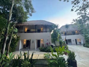 Un complejo turístico tropical en México con dos edificios blancos de techo de paja, rodeados de exuberante vegetación y palmeras. La cálida luz que emana de los balcones crea un ambiente sereno y acogedor, ideal para reuniones o conferencias al atardecer.