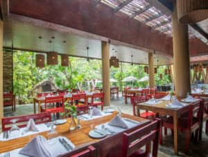Restaurante al aire libre con mesas de madera y sillas rojas, servilletas blancas y vasos. Ideal para reuniones o eventos, el espacio cuenta con grandes ventanales, vistas panorámicas, lámparas colgantes y plantas que crean un ambiente fresco y acogedor.