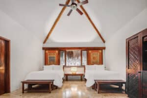 Una luminosa y minimalista habitación en uno de los mejores resorts de México cuenta con dos camas blancas, bancos de madera, un ventilador de techo, grandes ventanales con obras de arte estampadas en las paredes, un armario de madera a la derecha y elegantes suelos de baldosas de mármol claro, perfectos para eventos y conferencias.