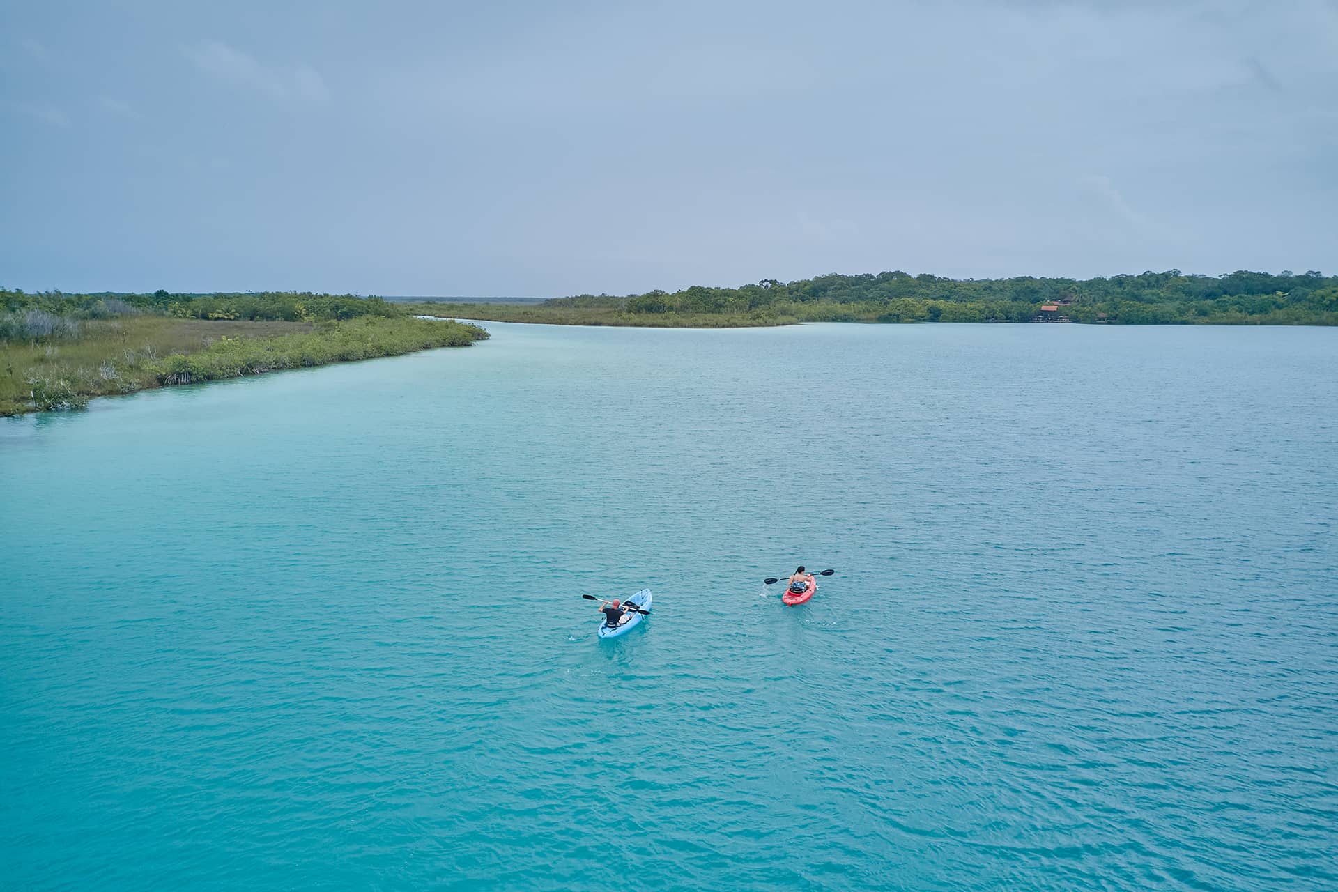 Two people kayak on clear turquoise water near a Conference Events Resorts Mexico property, surrounded by lush green vegetation under a cloudy sky. The blue and red kayaks add to the peaceful, scenic setting perfect for meetings or relaxation.