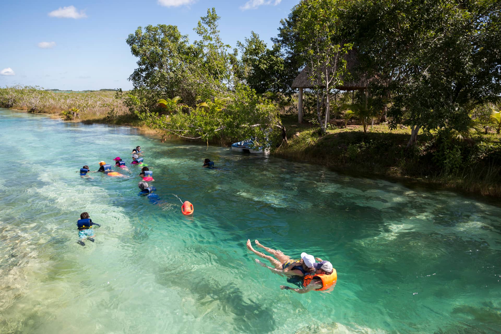 A group of people wearing life jackets float and swim in a clear, blue river at one of Mexico’s top resorts, surrounded by greenery and trees under a sunny sky. A thatched hut is visible on the bank.
