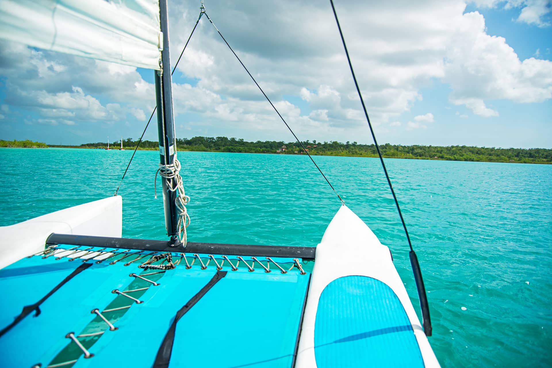View from a sailboat with blue netting and hulls, sailing on turquoise water toward a distant green shoreline, perfect for Mexico hotels or conference events under a partly cloudy sky.