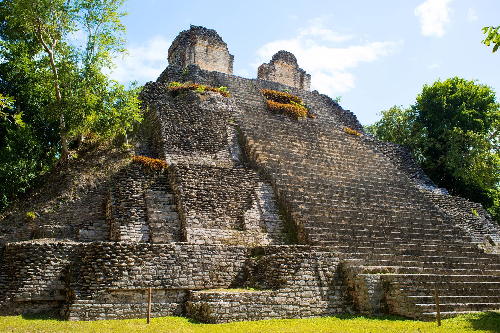 Ancient stone pyramid with steep steps and two structures on top, surrounded by lush green trees under a bright blue sky—discover this stunning landmark while staying at top Mexico hotels or attending conference events at nearby resorts.