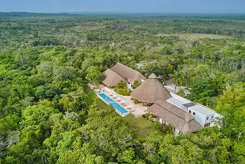 Aerial view of a resort with thatched-roof buildings and a long swimming pool, perfect for meetings and conference events, surrounded by dense green forest under a clear blue sky.