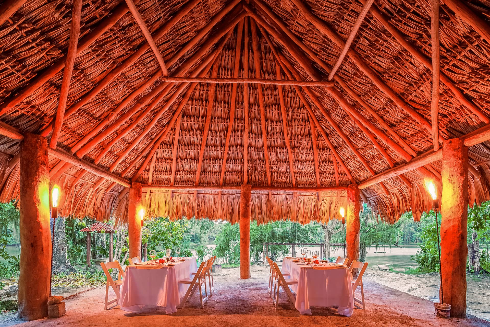 A rustic open-air pavilion with a thatched roof and wooden beams, perfect for intimate meetings or resort conference events in Mexico, featuring two tables set for dining amid lush greenery and softly lit by warm lamps.