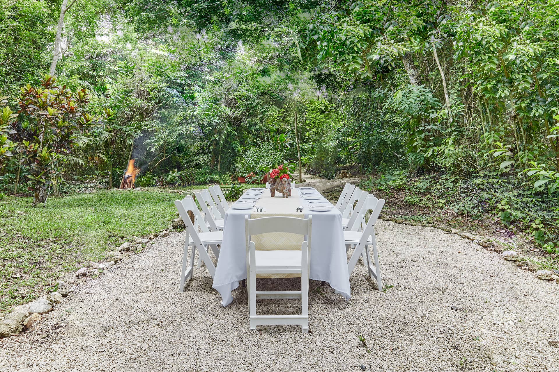 A long table with a white tablecloth and white chairs is set outdoors on a gravel path at one of Mexico’s luxurious resorts, surrounded by lush greenery. Red flowers adorn the table, while a small fire burns in the background—perfect for conference events or meetings.