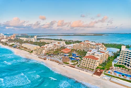Aerial view of a beachfront conference resort in Mexico with multiple pools, palm trees, and lounge chairs along a white sandy beach, adjacent to turquoise ocean waves under a partly cloudy sky. Perfect for meetings and events.