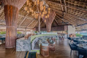 Un restaurante espacioso de estilo tropical con un alto techo de paja, columnas de madera, modernas lámparas de araña, asientos con cojines verdes y mesas de mármol; perfecto para eventos y conferencias en los resorts de México. Sus grandes ventanales ofrecen vistas al océano.