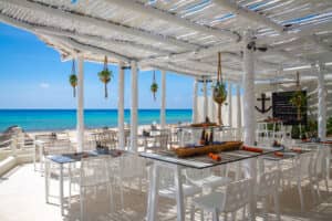 Restaurante al aire libre junto al mar, con mobiliario y decoración de madera blanca, a la sombra de una pérgola blanca. Las mesas ofrecen vistas al océano turquesa y al cielo azul, ideales para eventos o reuniones en los resorts mexicanos. Las plantas colgantes y los detalles náuticos realzan el ambiente costero.