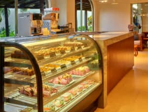 Una vitrina repleta de pasteles, tartas y postres variados se encuentra junto a un mostrador en una acogedora y bien iluminada cafetería de uno de los mejores hoteles de México, ofreciendo un lugar cálido para relajarse durante eventos o reuniones.