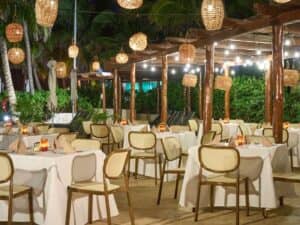 Restaurante al aire libre con pérgola de madera, lámparas colgantes de mimbre y guirnaldas de luces. Ideal para conferencias o reuniones, las mesas, vestidas con manteles blancos y velas, están rodeadas de vegetación y palmeras, creando un ambiente acogedor en los resorts de México.