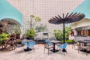 En los hoteles de México, una colorida cafetería al aire libre cuenta con sillas azules y negras, mesas negras, una gran sombrilla artística, exuberantes plantas verdes, una jardinera con forma de rueda de carreta y una pared blanca estampada, perfecta para reuniones informales o eventos de conferencias.