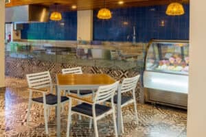 El interior de una cafetería moderna en un complejo turístico de México, con suelo de baldosas estampadas, una mesa de madera con cuatro sillas blancas, una pared de azulejos azules, luces colgantes y una vitrina con helados de colores, perfecta para reuniones informales o eventos de conferencias.
