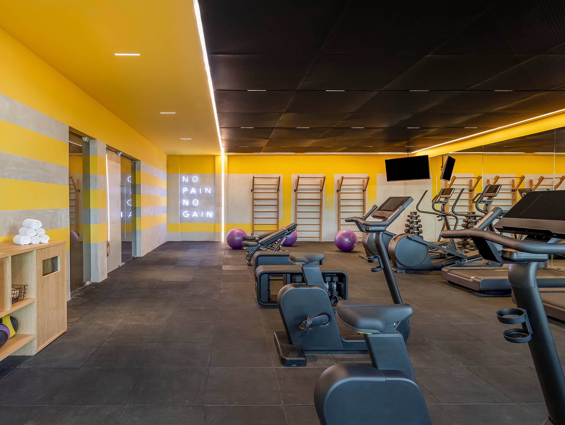 Modern gym at our Mexico resort with yellow walls, black ceiling, cardio machines, bikes, purple exercise balls, wall bars, and a large mirror. A neon NO PAIN NO GAIN sign inspires guests during meetings or conference events. Towels are stacked neatly on a shelf.