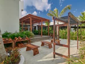 Zona de estar al aire libre con bancos de madera, mesas y pérgolas rodeada de exuberantes plantas verdes y palmeras sobre arena clara; perfecta para eventos o reuniones en complejos turísticos de México cerca de un edificio moderno bajo un cielo azul con nubes.