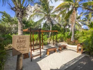 Gimnasio al aire libre sobre arena, rodeado de palmeras y plantas tropicales; con equipos y bancos de madera. Ideal para huéspedes de hoteles en México o para eventos de congresos. Un cartel de madera reza: «Entrena duro, mantente fresco con el hashtag #FACondesa».
