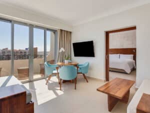 Una luminosa suite de hotel ideal para reuniones, con sala de estar, mesa redonda de madera con cuatro sillas azules, televisor de pantalla plana y un dormitorio con una cama grande. Las puertas de cristal dan a un balcón con mobiliario de exterior y vistas a la ciudad.