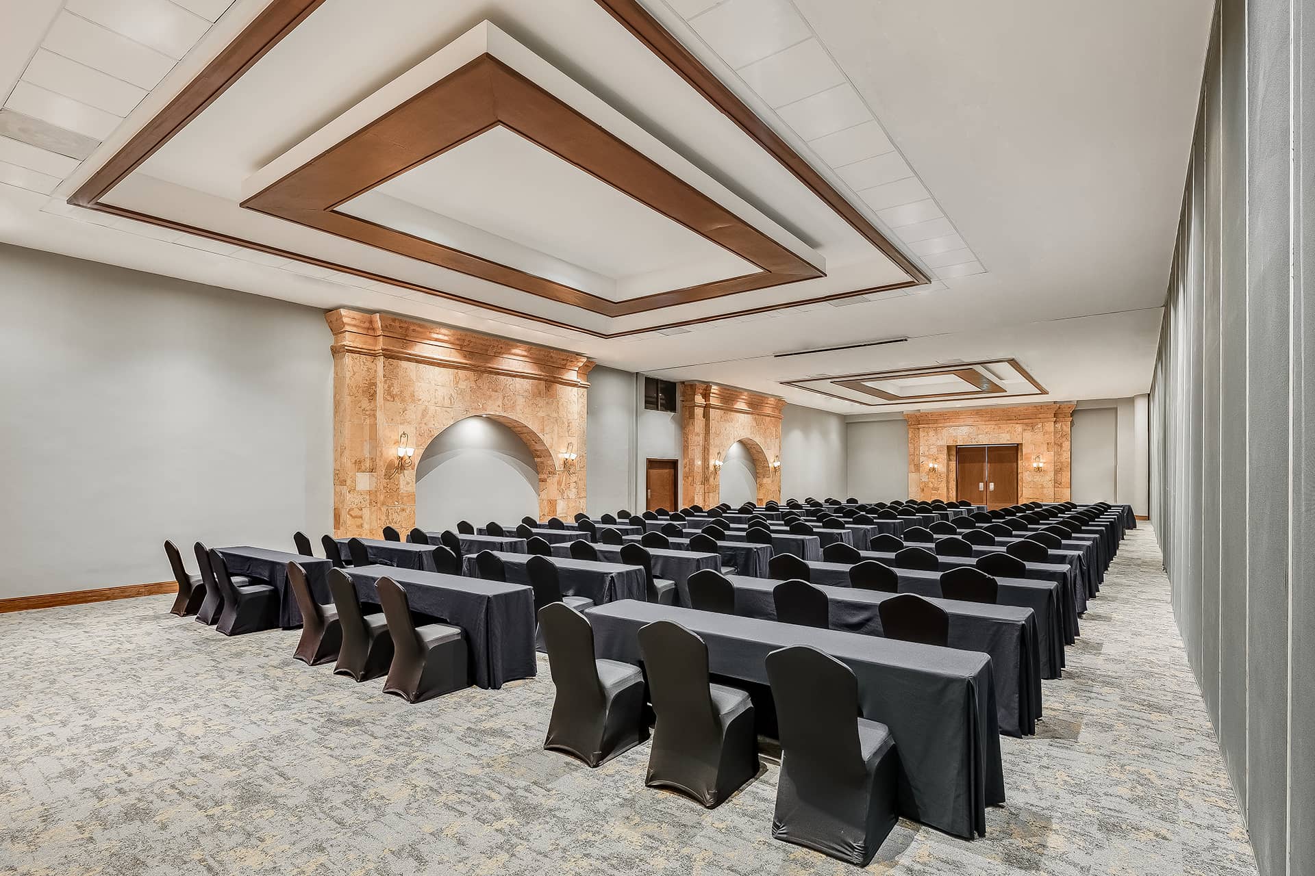 A spacious conference room at one of Mexico’s top resorts, with rows of black-covered tables and chairs, elegant lighting, marble walls, beige carpet, and a modern geometric ceiling—perfect for formal meetings or conference events.