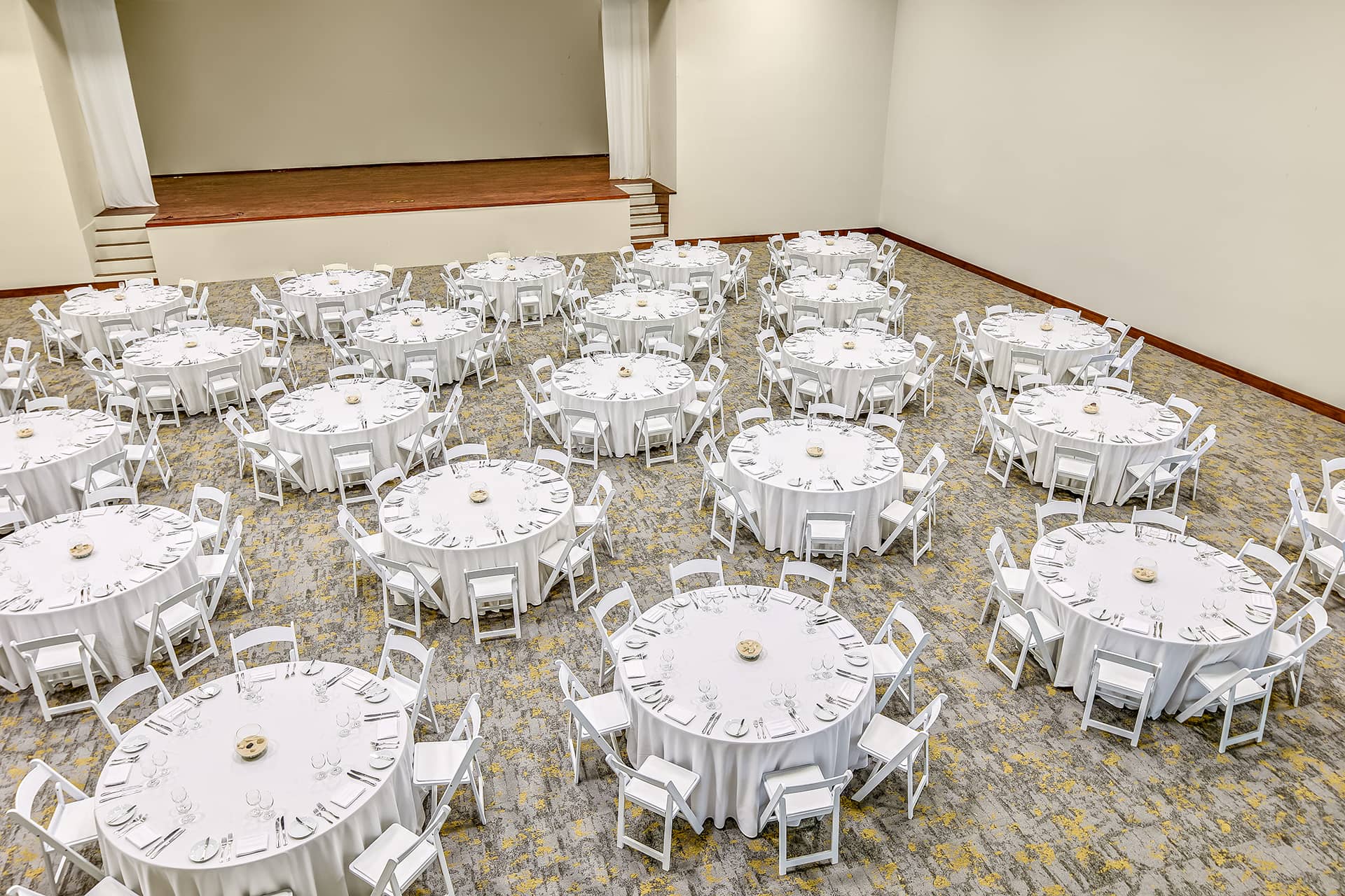 A large banquet hall at one of Mexico's top resorts, with round tables covered in white tablecloths, each set for meetings and conference events. The tables are evenly spaced, and there is a stage at the front of the room.