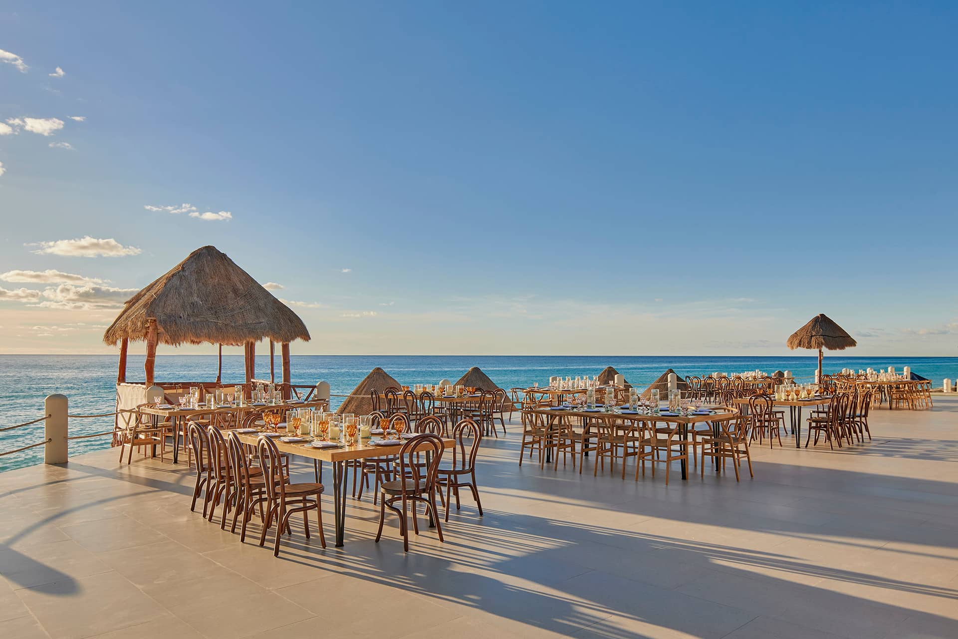 Outdoor dining area with wooden tables and chairs, some under thatched-roof palapas, overlooks a calm blue ocean—an inviting setting at one of Mexico’s top resorts, perfect for conference events or meetings under a clear sky.