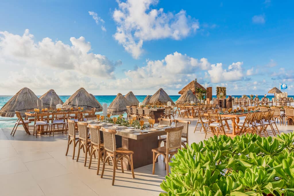 Outdoor beachfront dining area at a Mexico resort, with wooden tables and chairs set for conference events or meetings. Palapa-style straw huts and lush greenery create a scenic setting under a bright blue sky overlooking the ocean.