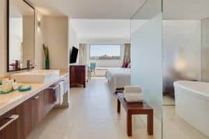 Suite de hotel moderna, perfecta para eventos y conferencias, con un amplio baño que incluye dos lavabos, bañera y banco de madera. El dormitorio cuenta con una cama king size, TV y una gran ventana con impresionantes vistas a la ciudad, ideal para hoteles y reuniones en México.