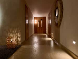 Un pasillo con iluminación cálida en los complejos turísticos de México, con paredes de color beige, puertas de madera a cada lado, una lámpara de mimbre que proyecta sombras, apliques de pared al final y un espejo circular con marco de fibra natural en la pared derecha: perfecto para eventos o reuniones.