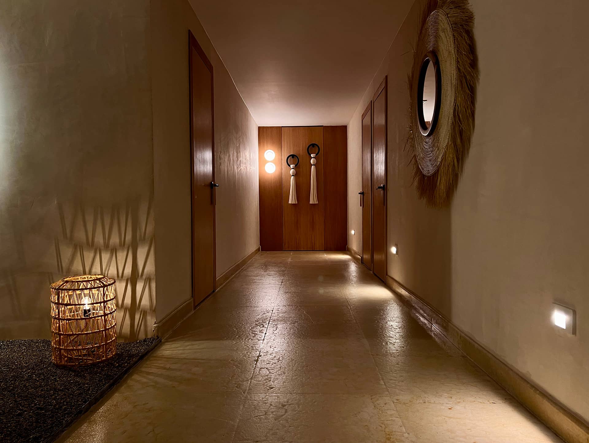 A warmly lit hallway in Mexico resorts, with beige walls, wooden doors on each side, a woven basket lamp casting shadows, wall sconces at the end, and a circular mirror with a natural fiber frame on the right wall—perfect for conference events or meetings.