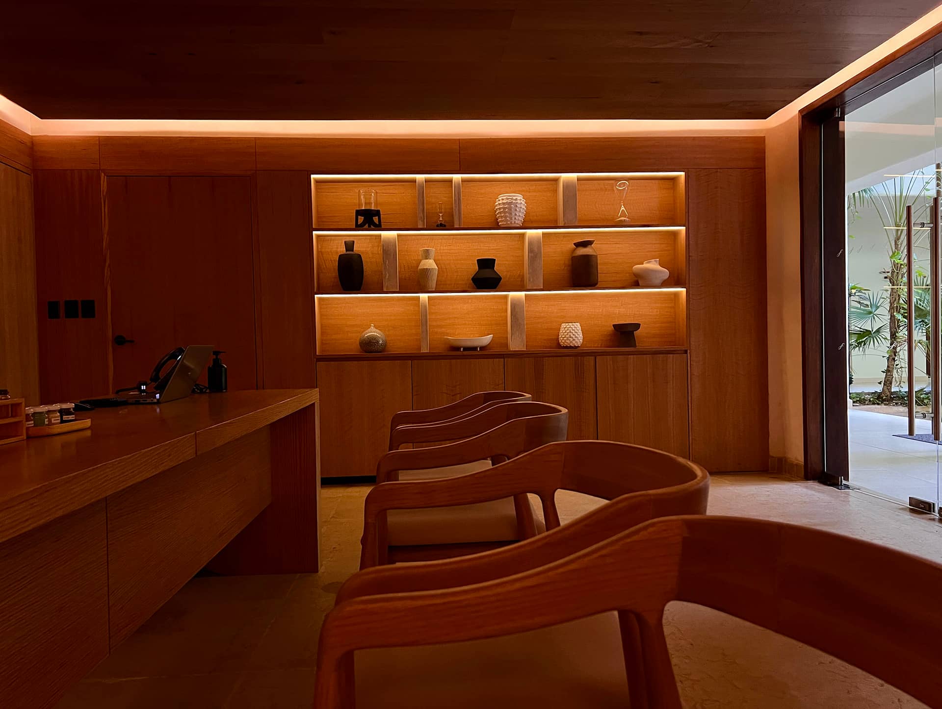 A modern, warmly lit office with wooden chairs and desk, built-in shelves displaying various pottery and vases, perfect for small meetings, and a large window revealing plants outside.