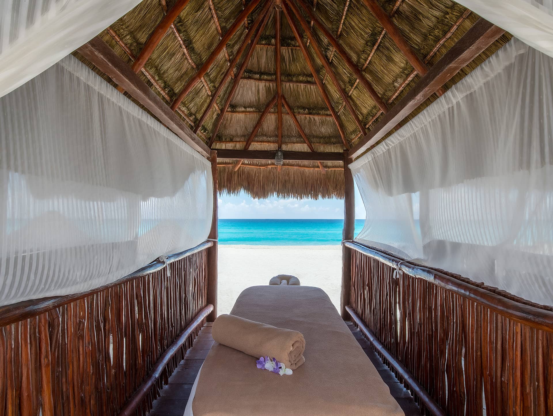 A massage table with a rolled towel and flowers sits under a thatched-roof cabana on a sandy beach at one of Mexico’s premier resorts, facing the ocean with clear blue water and sky in the background.