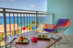 Un balcón con vistas al océano en uno de los resorts más exclusivos de México, con una mesa puesta para dos personas que incluye fruta, aperitivos y coloridos cócteles. Una moderna silla multicolor completa la escena, ideal para relajarse después de eventos o reuniones.