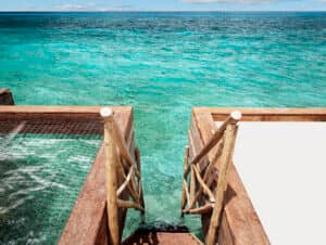 Una escalera de madera con barandillas rústicas desciende desde una terraza en uno de los mejores complejos turísticos de México hasta las cristalinas aguas turquesas del océano, con un recinto de redes de pesca a la izquierda y el mar abierto extendiéndose hasta el horizonte bajo un cielo parcialmente nublado.