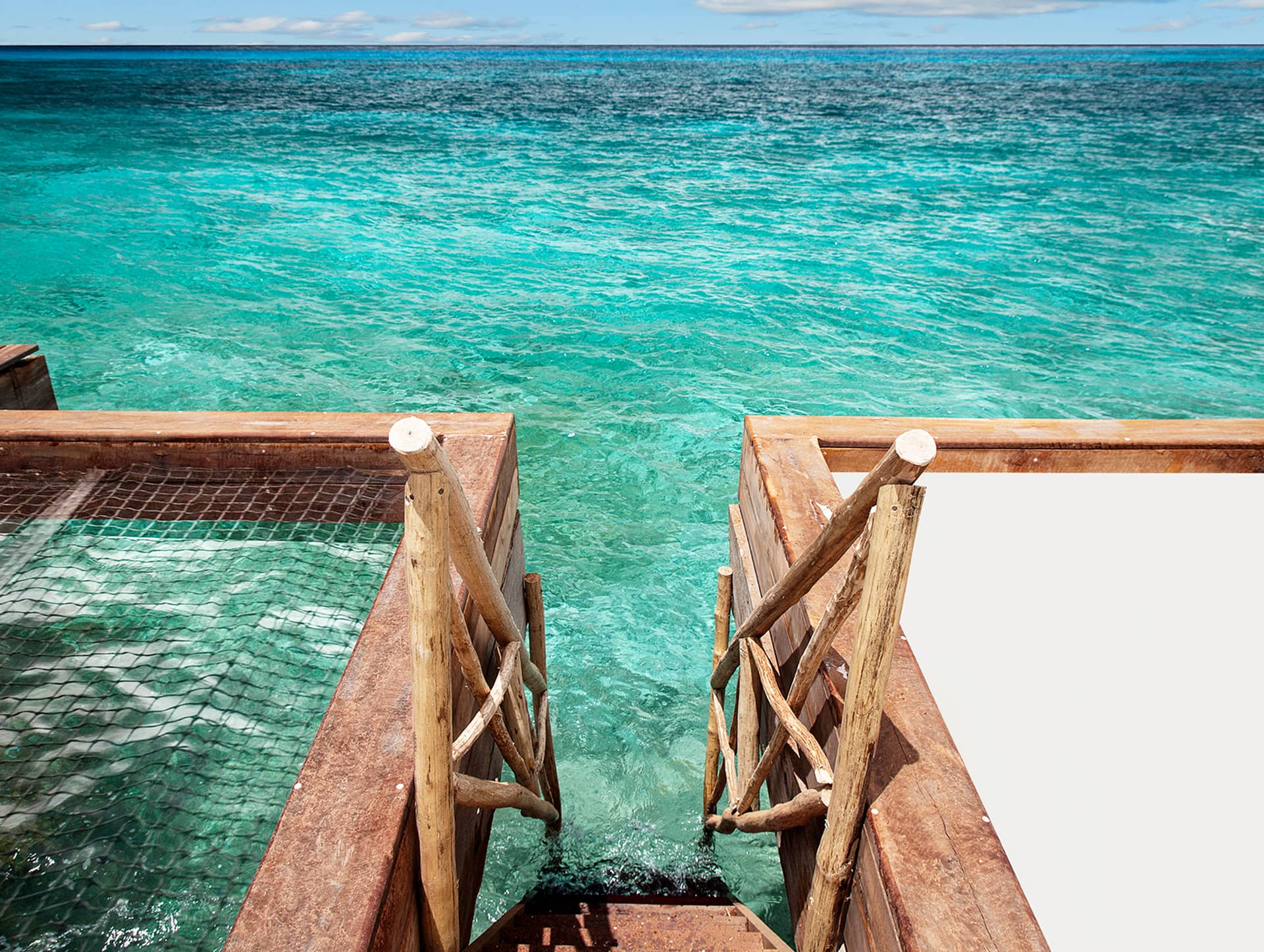 A wooden staircase with rustic railings leads down from a deck at one of Mexico’s finest resorts into clear turquoise ocean water, with a fishnet enclosure on the left and open sea stretching to the horizon under a partly cloudy sky.