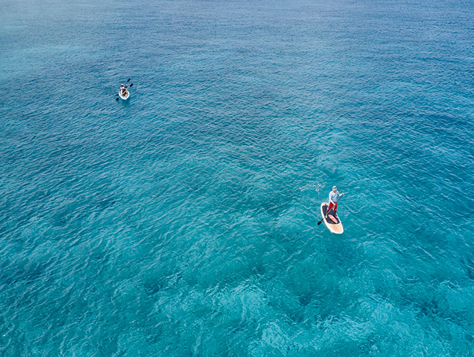 Aerial view of two people on clear blue water at one of Mexico’s top resorts; one person is paddleboarding in the foreground, while another is kayaking farther away in the background. Perfect for conference events and meetings.