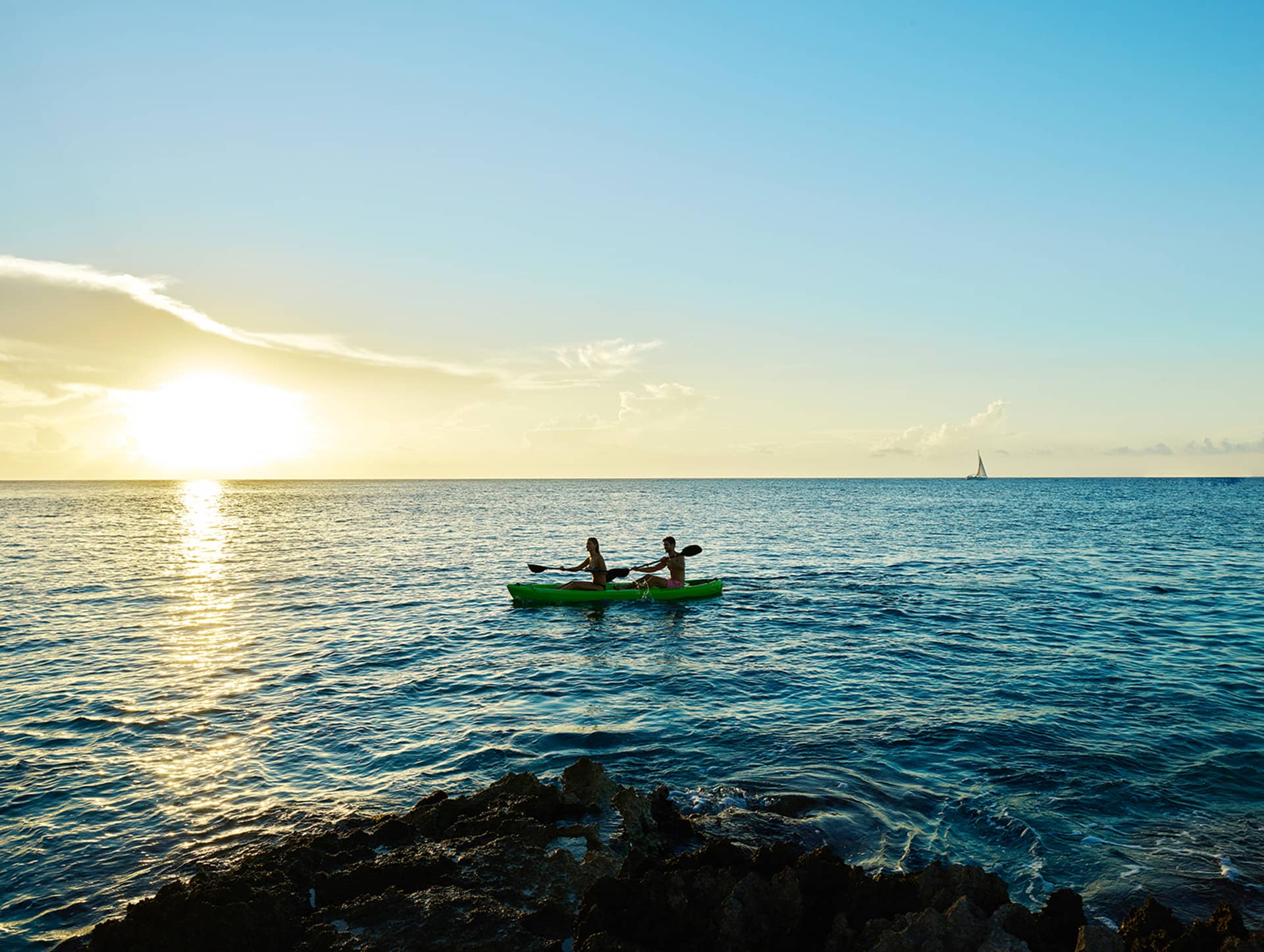 Two people kayaking on calm blue water at sunset near a Mexico resort, with a sailboat in the distance and rocky shoreline in the foreground. The sky is mostly clear with a few clouds and the sun low on the horizon.