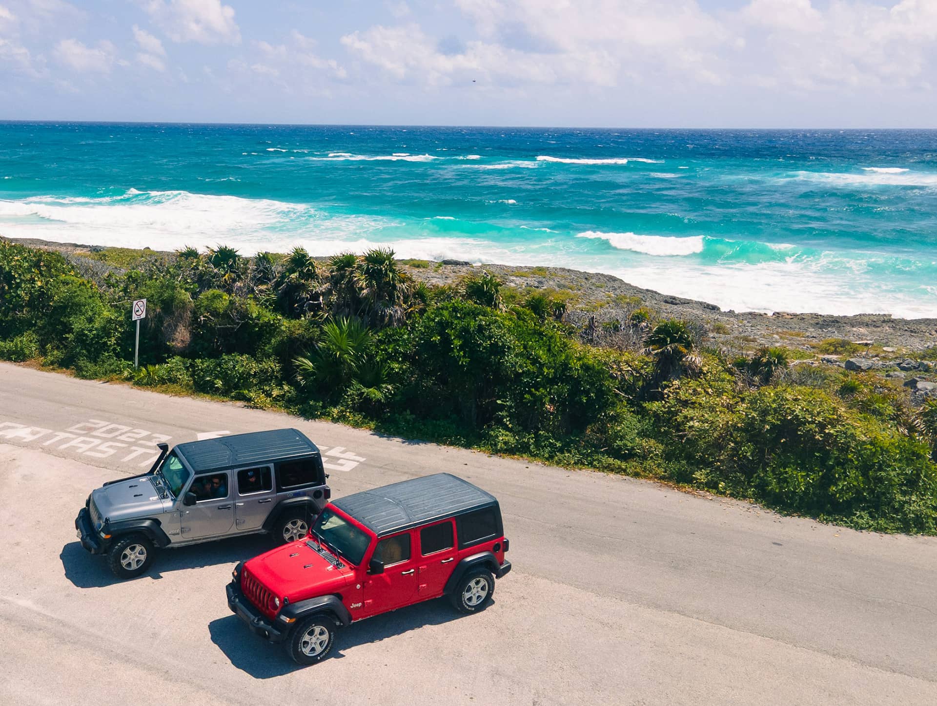 Two Jeeps, one gray and one red, are parked on a coastal road next to lush greenery and rocky terrain, with turquoise ocean waves and a partly cloudy sky—perfect for exploring near Mexico hotels or conference events resorts.