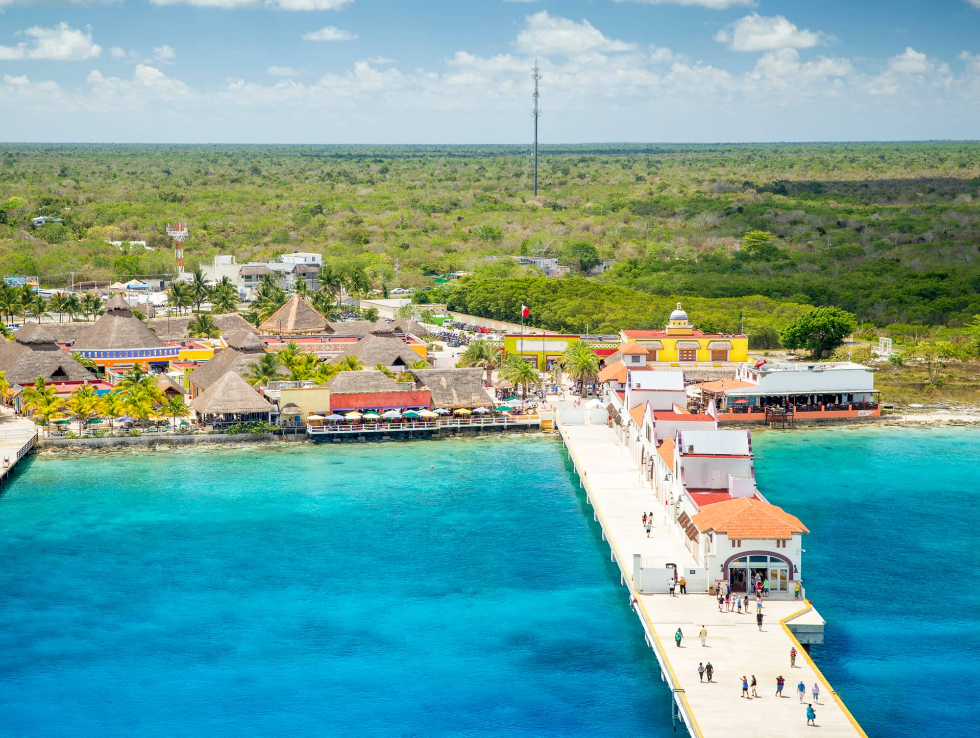 A long pier extends over turquoise water toward a lively coastal village with colorful buildings, palm trees, and greenery—an inviting scene reminiscent of conference events at Mexico resorts set against a lush forest and partly cloudy sky.