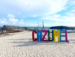 Un colorido letrero de COZUMEL se alza sobre una playa de arena con rocas dispersas, cielo azul y olas del mar de fondo. Ideal para conferencias o reuniones en hoteles y resorts cercanos de México, con madera flotante decorativa detrás del letrero.